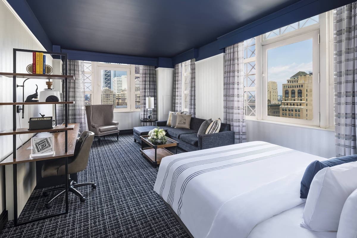 Une chambre d’hôtel avec un grand lit, un bureau avec une chaise, un canapé et une vue sur le paysage urbain grâce à de grandes fenêtres.