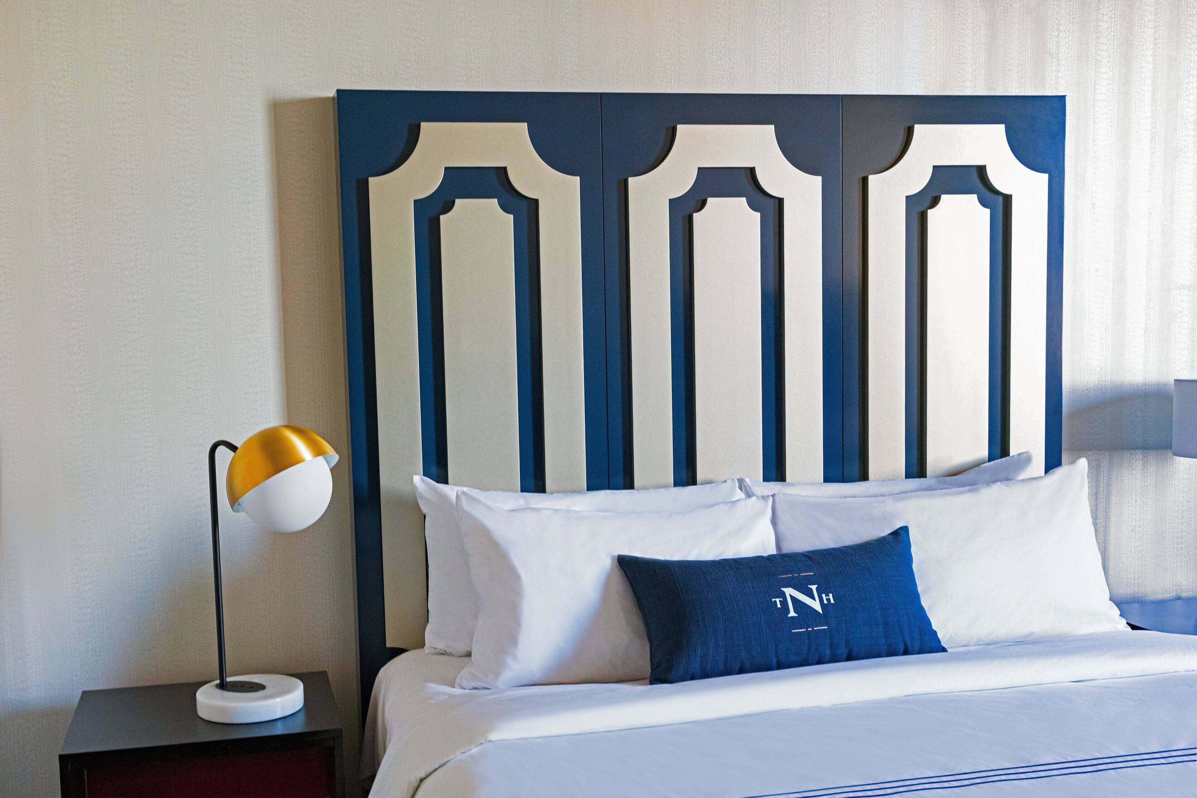 Quarto de hotel com cama, mesa de cabeceira, abajur e cabeceira com linhas verticais.