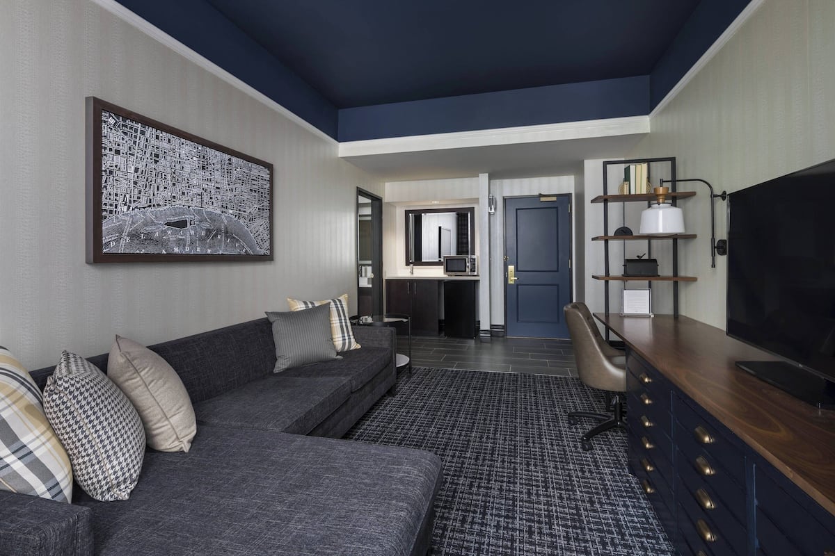 Une chambre d’hôtel moderne avec un canapé gris, une grande œuvre d’art représentant un paysage urbain en noir et blanc, un téléviseur à écran plat, un bureau en bois et un plafond bleu foncé.
