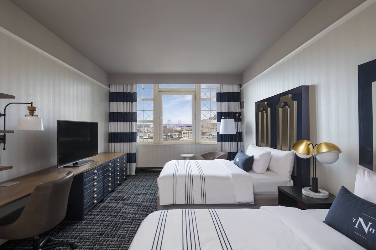 Une chambre d’hôtel comprenant un lit, un bureau, une télévision, une commode, une fenêtre avec des rideaux et une table de chevet avec une lampe.