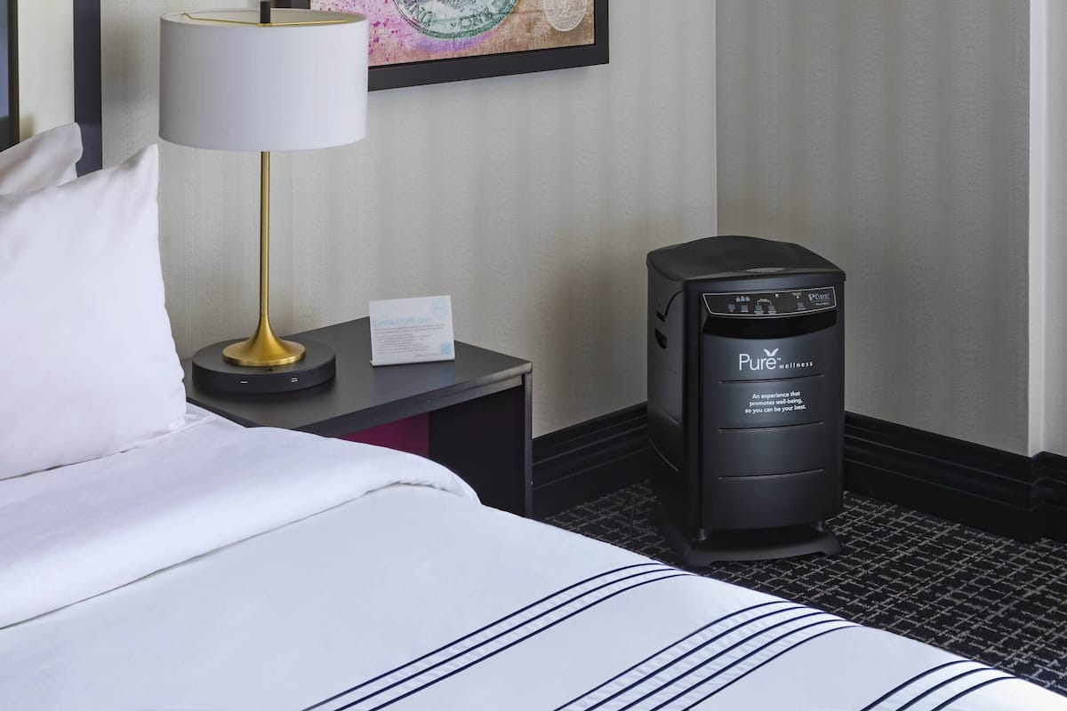 Une chambre d’hôtel équipée d’un lit, d’un bureau, d’une lampe et d’un haut-parleur noir portant l’inscription « Pure Wellness ».