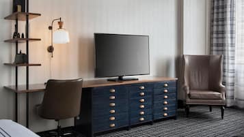 Habitación de hotel con televisor de pantalla plana sobre un mueble de madera, un sillón de cuero, un escritorio con silla y una estantería de pared con objetos decorativos.