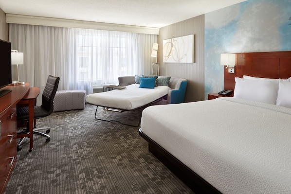 A hotel room with a large bed, a smaller bed, a desk with a chair, a sofa, and a TV.