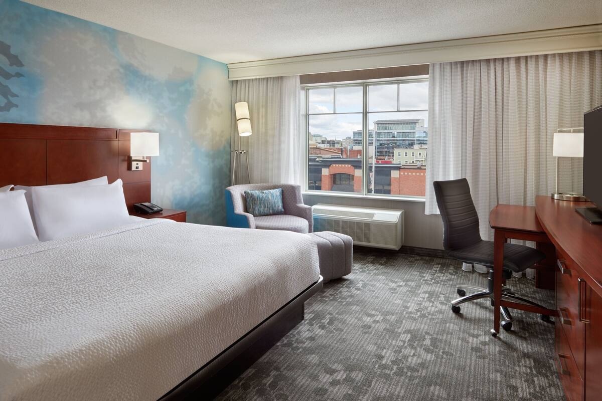 Habitación de hotel con una cama grande, un escritorio, una silla y vista a la ciudad por la ventana.