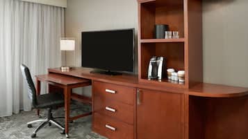 A hotel room with a wooden desk, a flat-screen TV, a lamp, a coffee maker, and a cup holder.