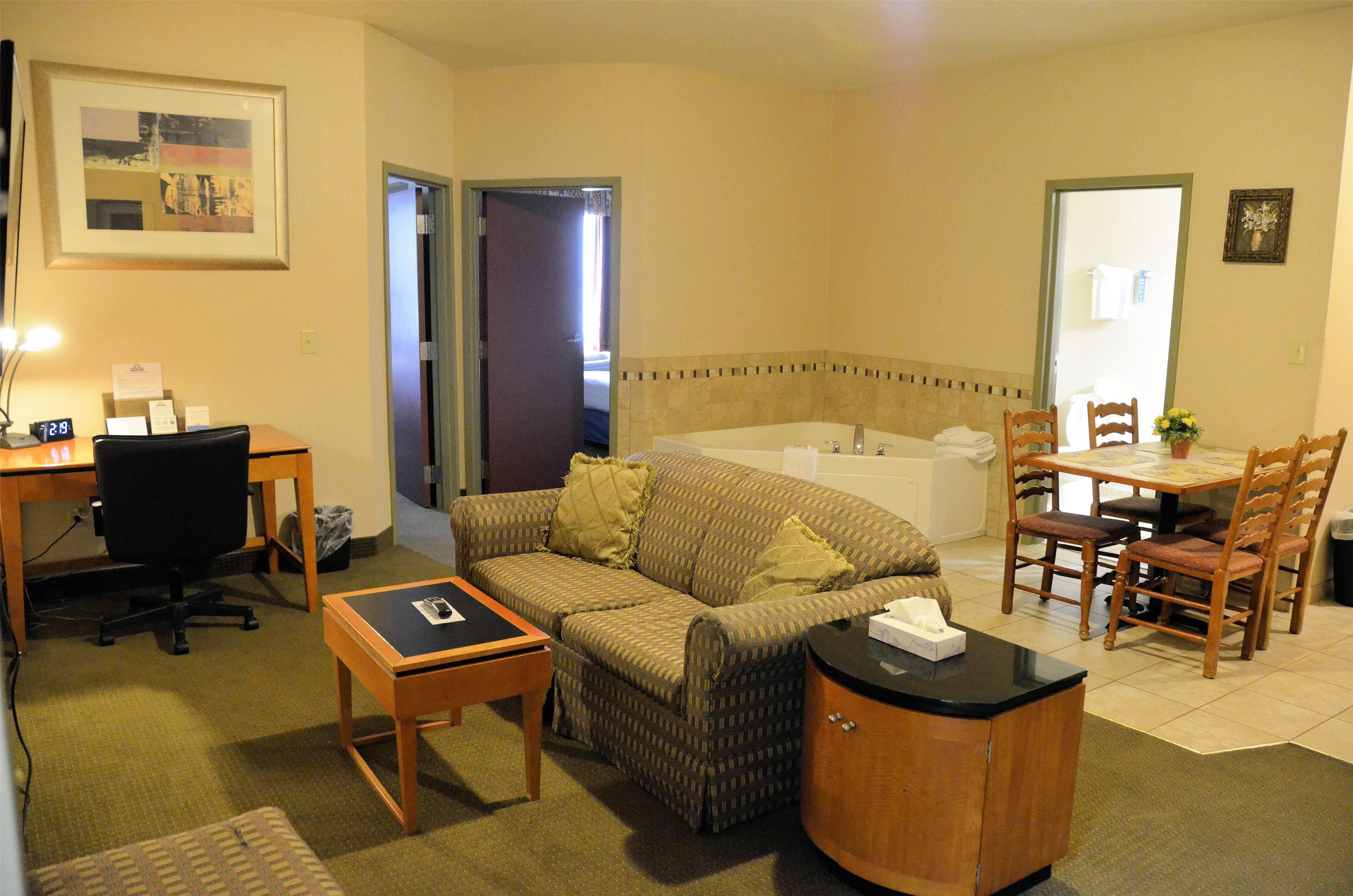 A hotel room with a sofa, a desk with a computer, a small table, a cabinet, a dining table with chairs, and a bathroom with a bathtub.