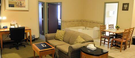 A hotel room with a sofa, a desk with a computer, a small table, a cabinet, a dining table with chairs, and a bathroom with a bathtub.
