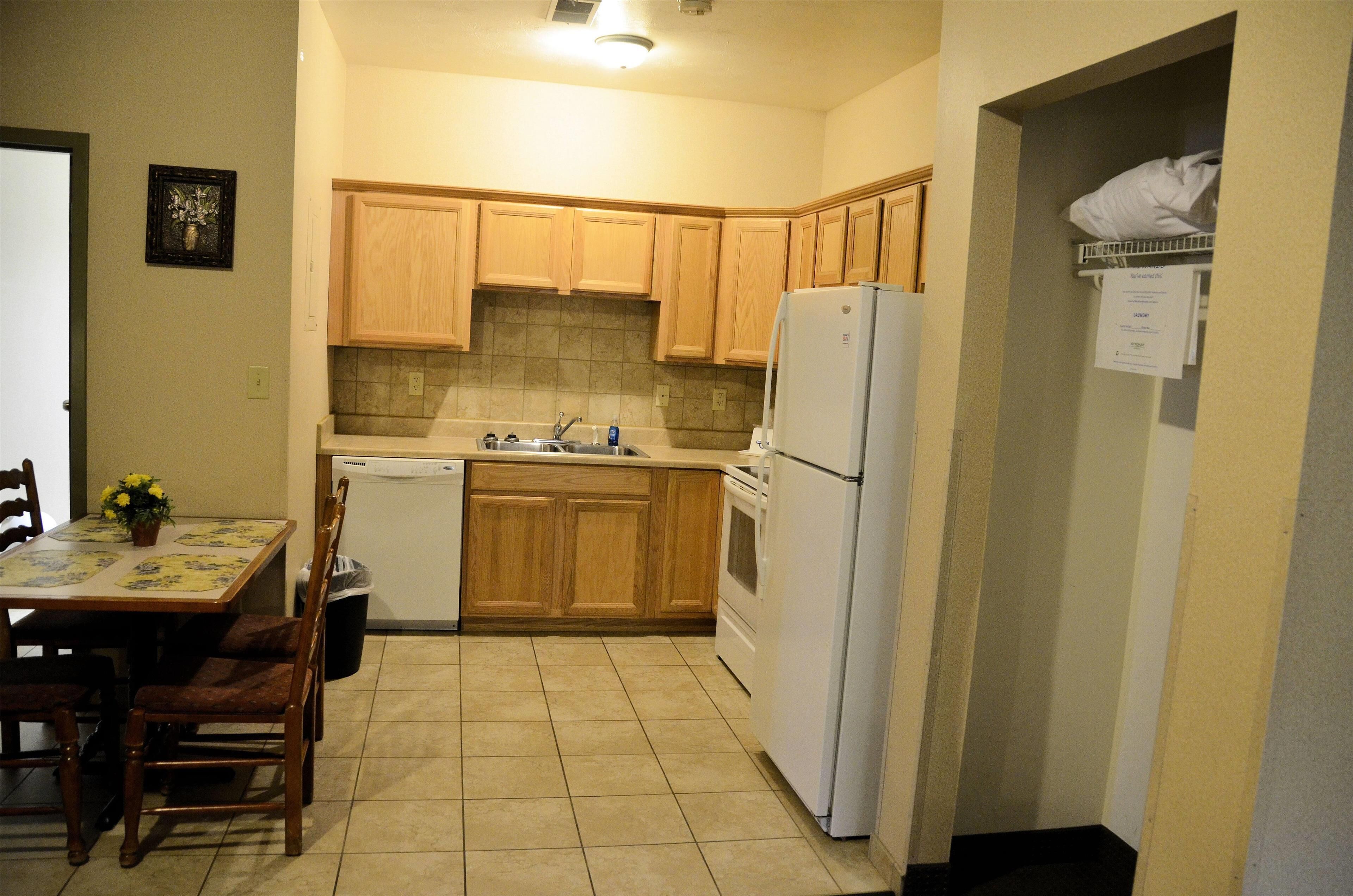 A compact kitchen with wooden cabinets, a dishwasher, a refrigerator, and a dining table with chairs.