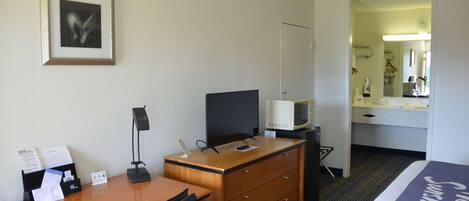 A hotel room with a bed, a desk with a lamp, a television, a dresser, and a framed picture on the wall.