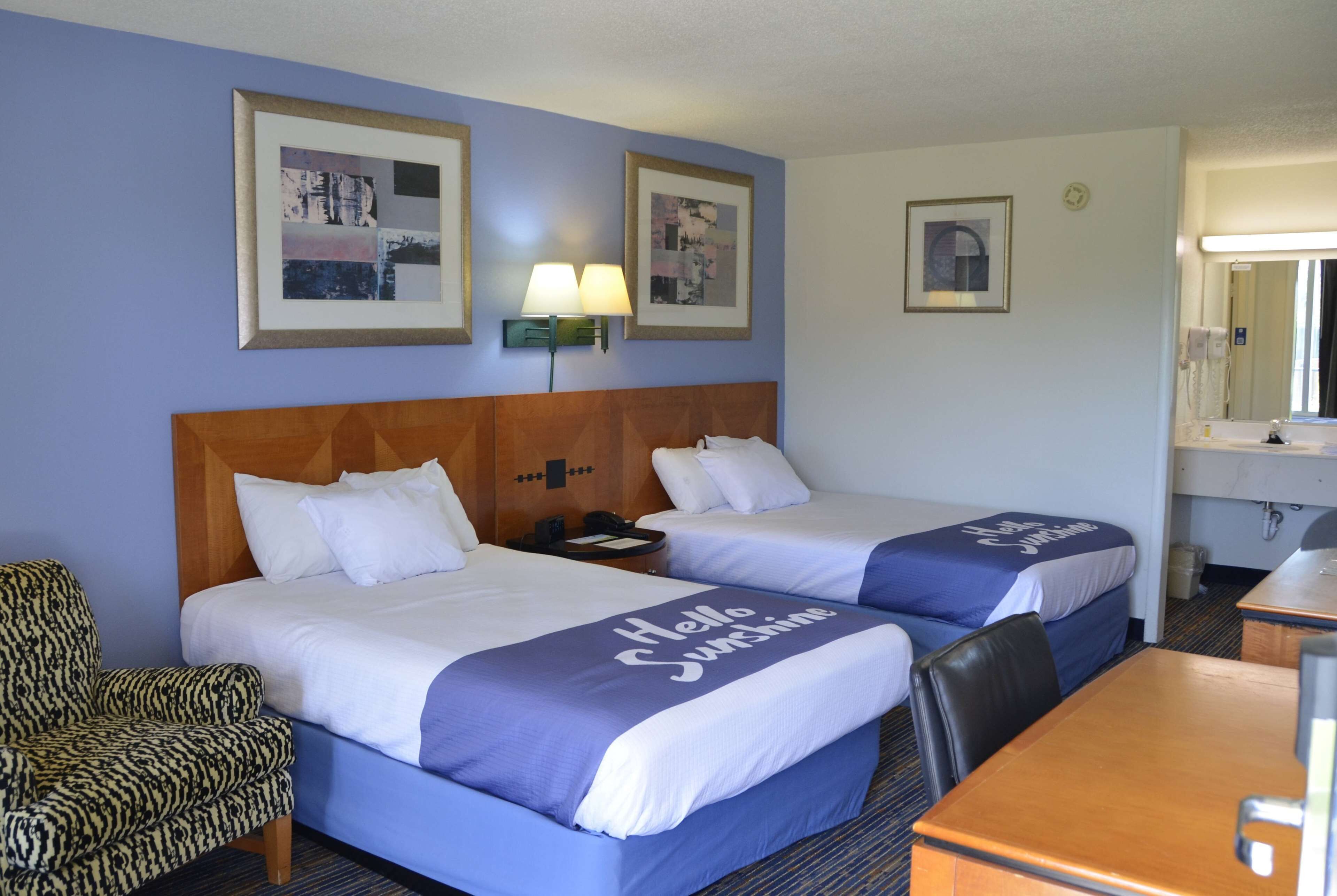 A hotel room with two beds, a wooden headboard, a chair, a desk, and framed pictures on the wall.