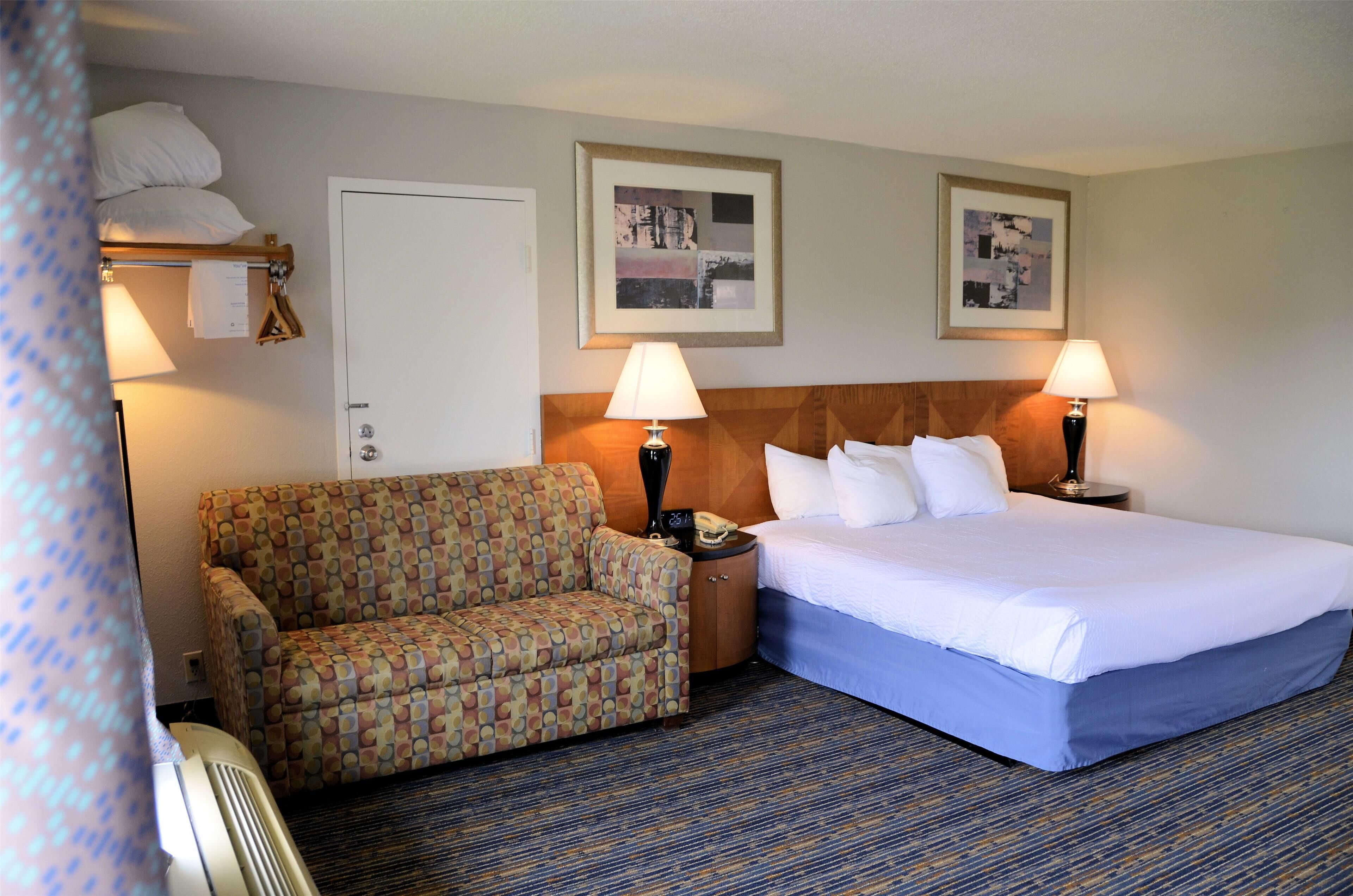 A hotel room with a bed, a patterned armchair, two bedside lamps, and two framed pictures on the wall.