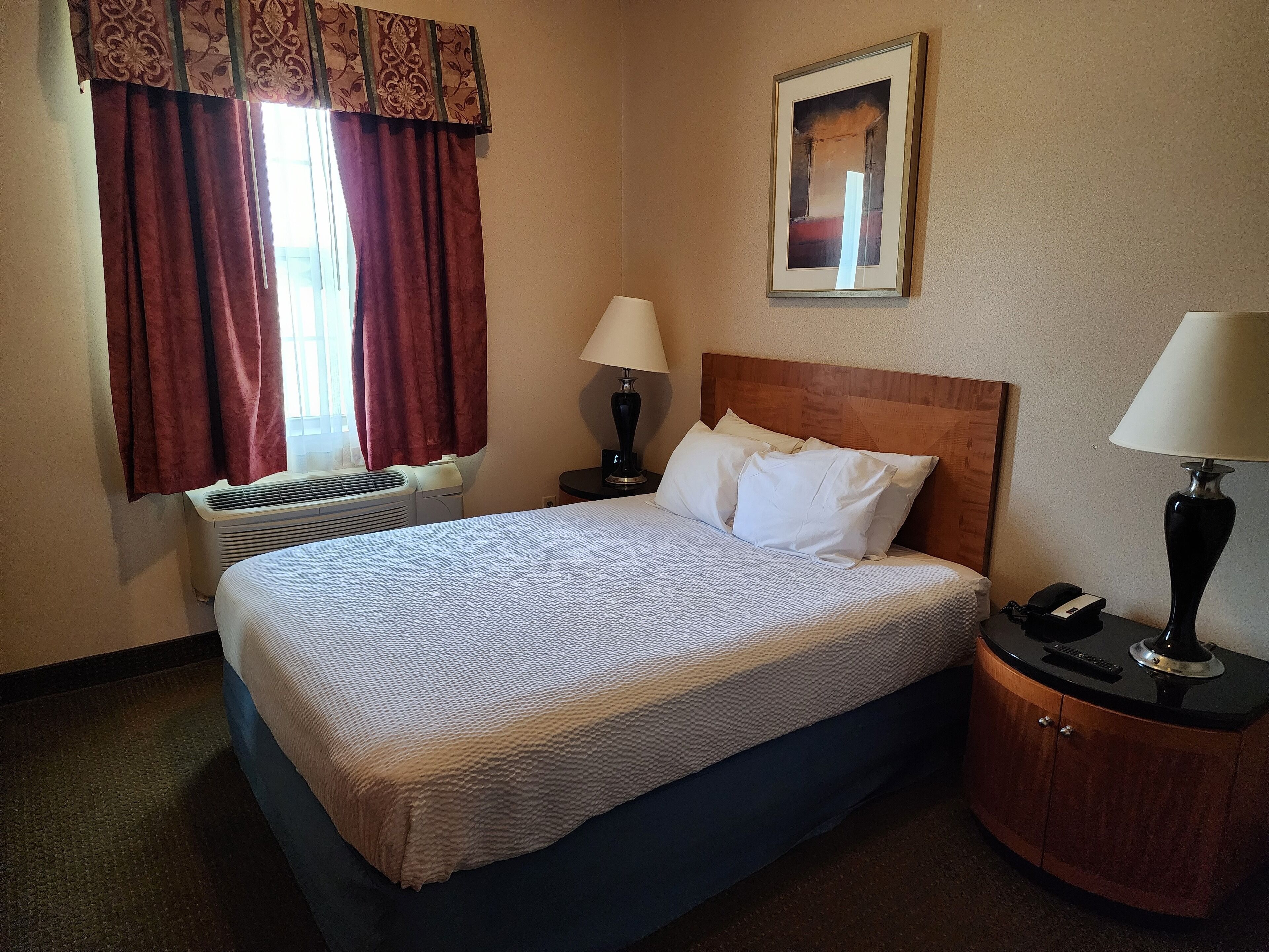 A hotel room with a bed, two bedside tables, a wall-mounted lamp, a framed picture, and a window with curtains.