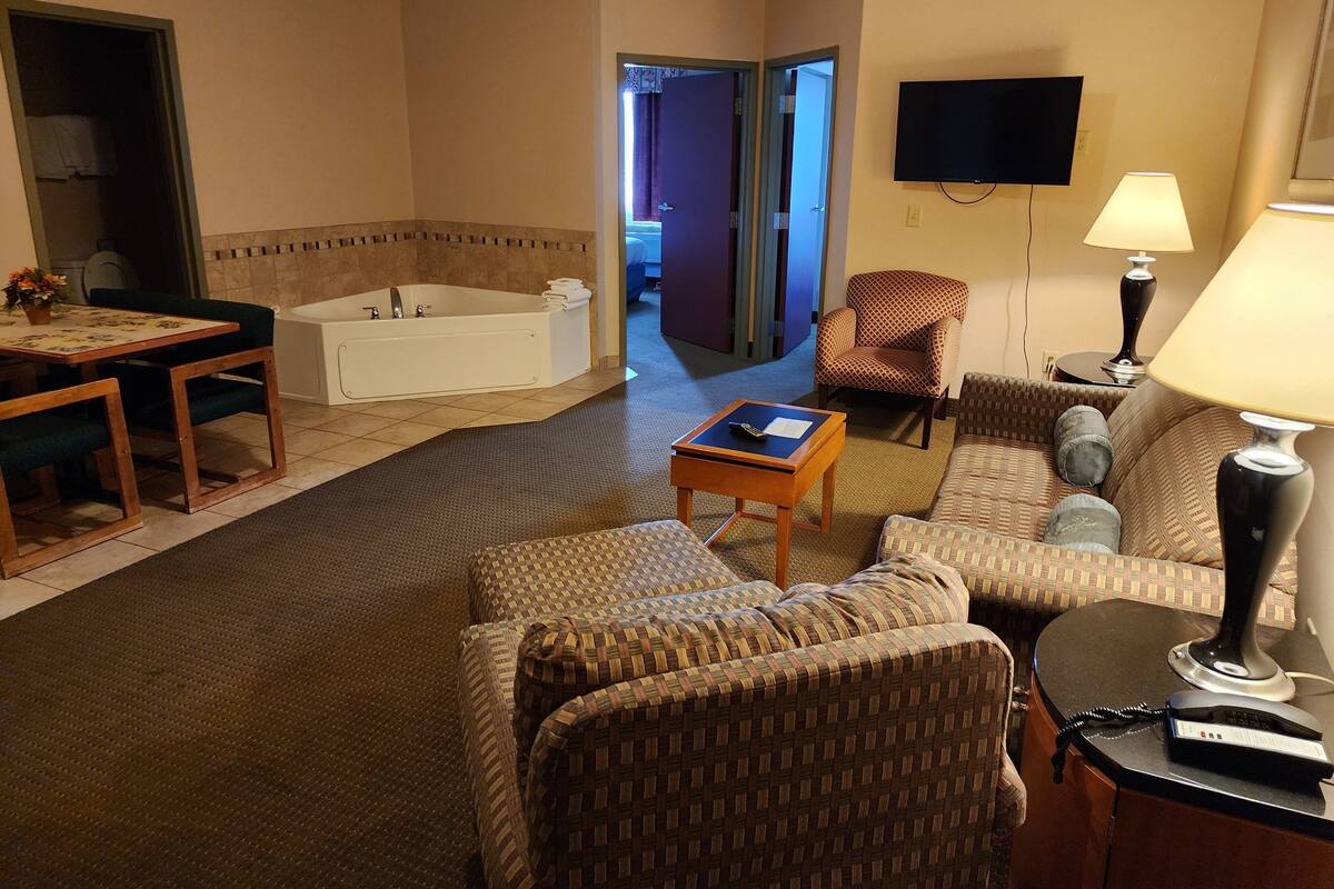 A hotel room with a living area, a dining table, a sofa, a chair, a TV, a lamp, and a bathtub.