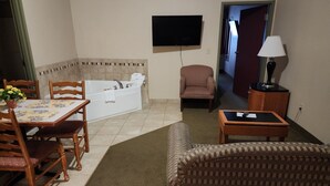 Hotellihuone, jossa on ruokailutila, sohva, tuoli, pieni pöytä, taulutelevisio, lamppu ja kylpyamme.