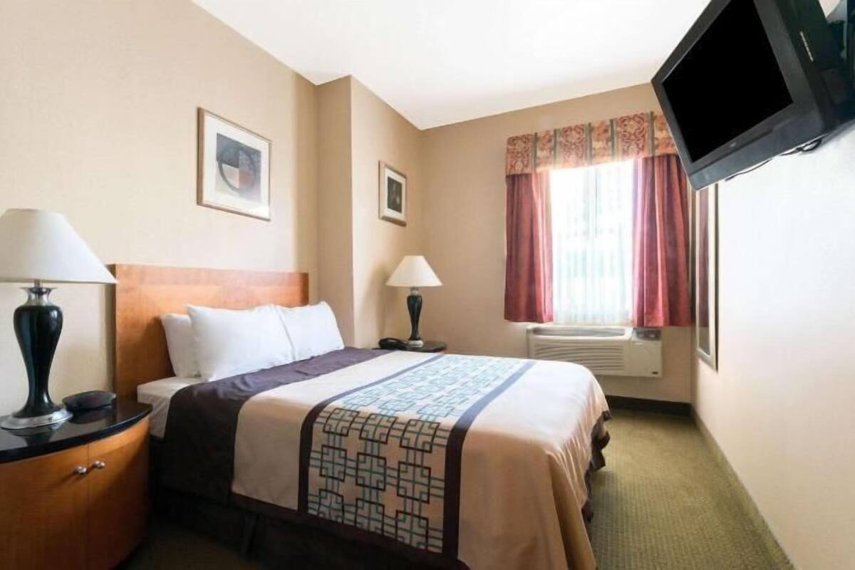 A hotel room with a bed, two bedside tables with lamps, a television mounted on the wall, and a window with curtains.