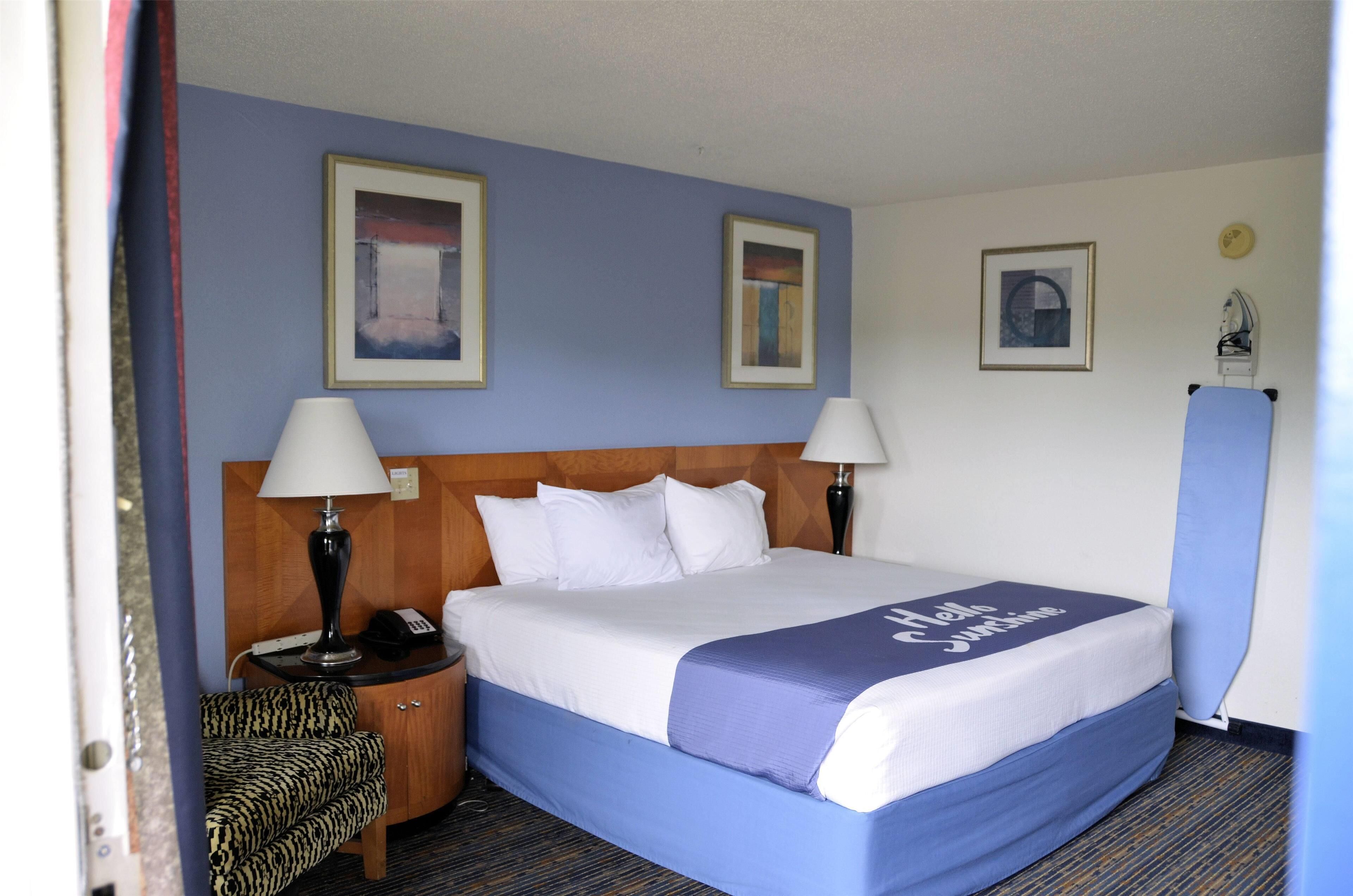 Habitación de hotel con cama, dos lámparas de noche, mesita de noche, una silla y dos cuadros en la pared.