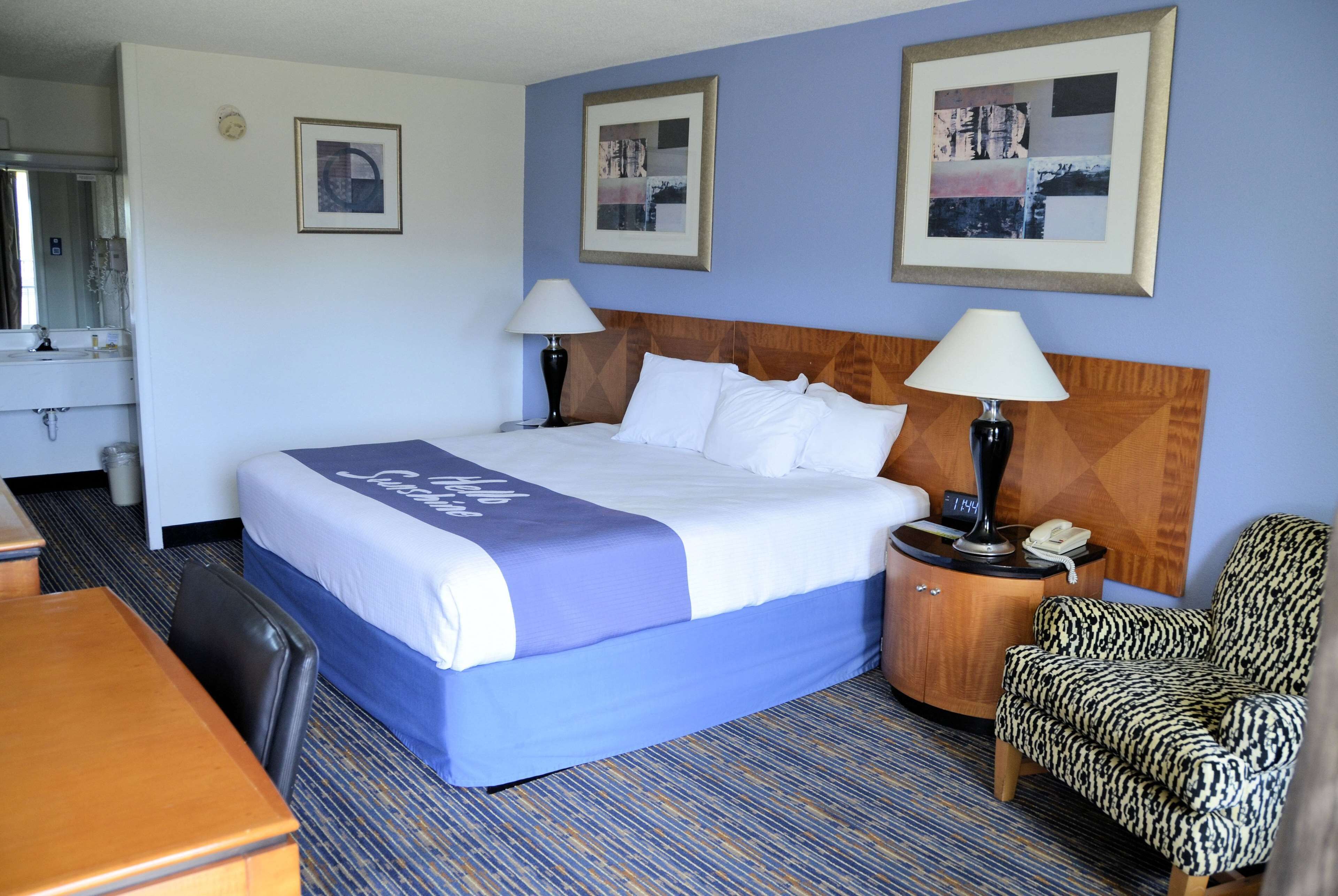 A hotel room with a large bed, two bedside lamps, a wooden nightstand, a patterned armchair, and a desk with a chair.