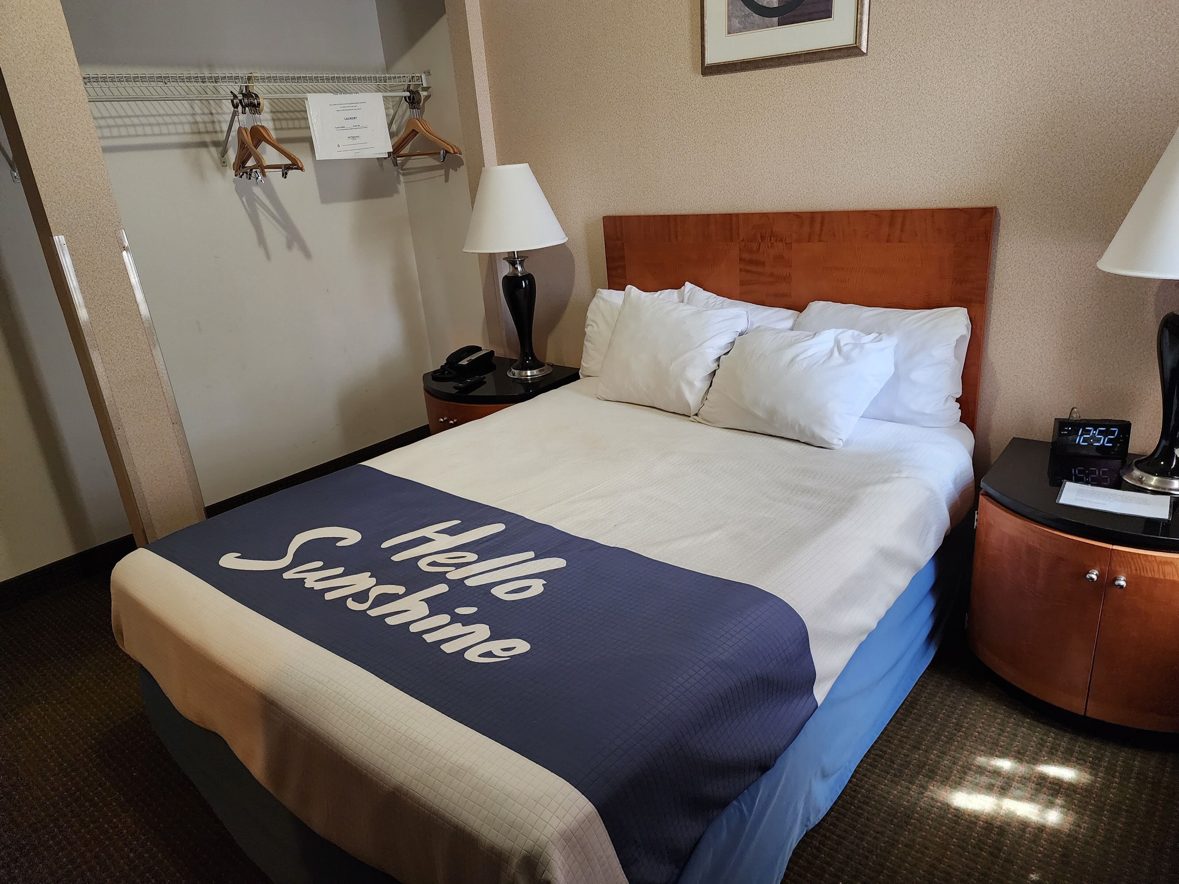 A hotel room with a bed, two bedside lamps, a nightstand with a phone, a digital clock, and a welcome sign on the bed.