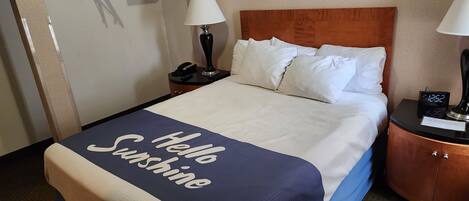A hotel room with a bed, two bedside lamps, a nightstand with a phone, a digital clock, and a welcome sign on the bed.