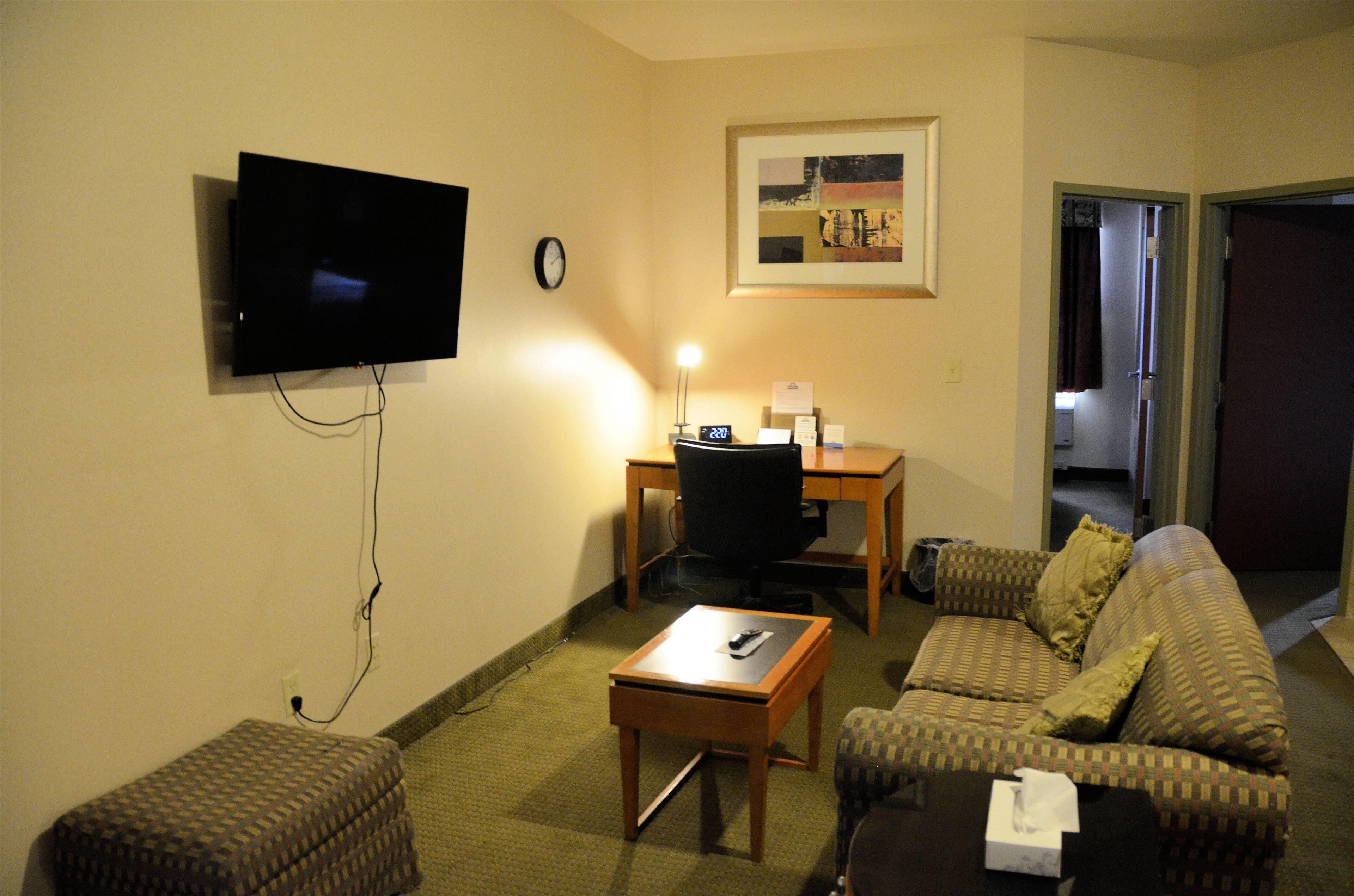 A small living room with a flat-screen TV, a desk, a chair, a sofa, and a small ottoman.