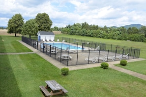 A fenced swimming pool area with lounge chairs and a small building, surrounded by well-maintained grass and trees.