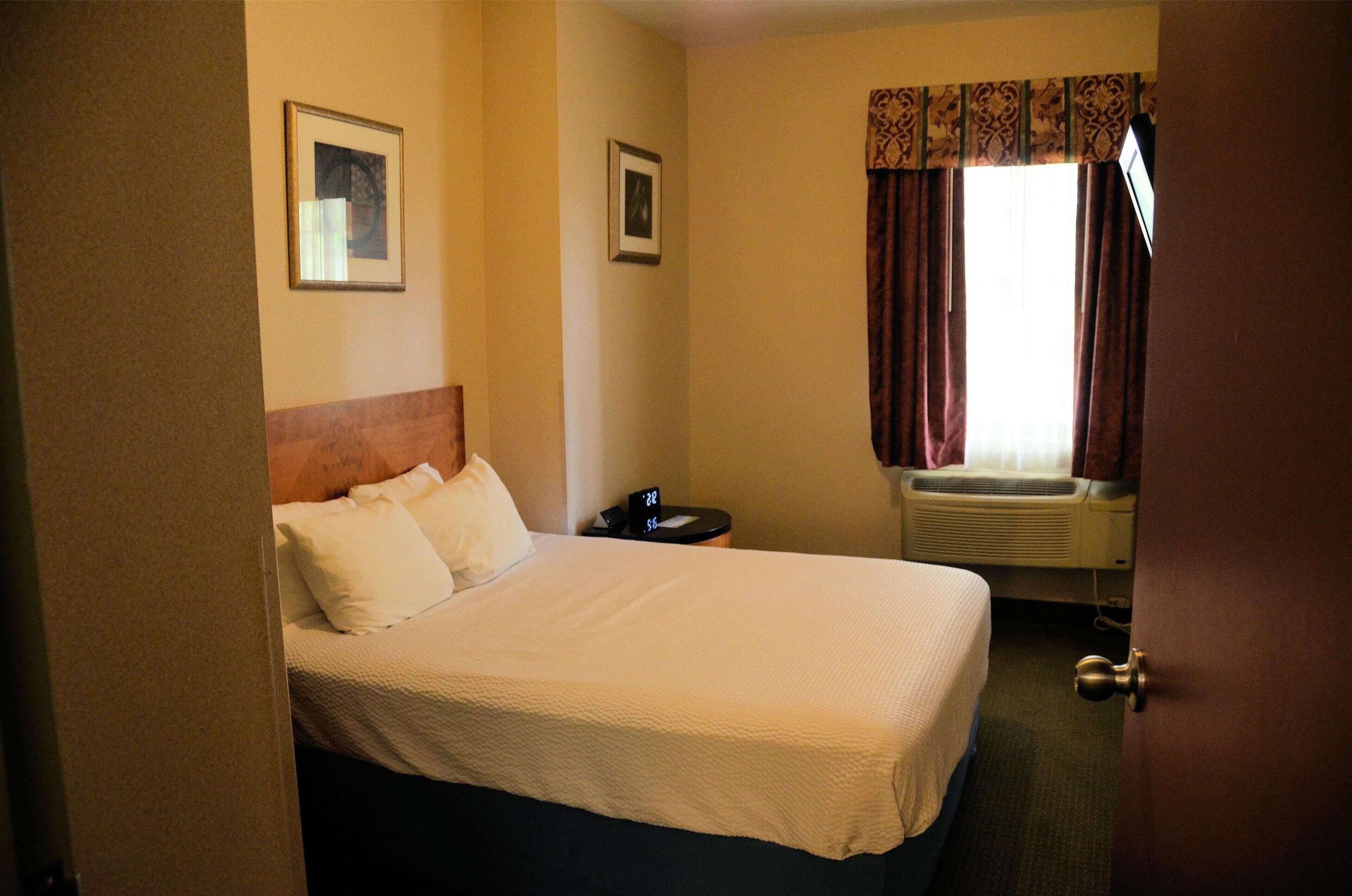 A hotel room with a bed, a nightstand, a window with curtains, and a wall-mounted air conditioning unit.