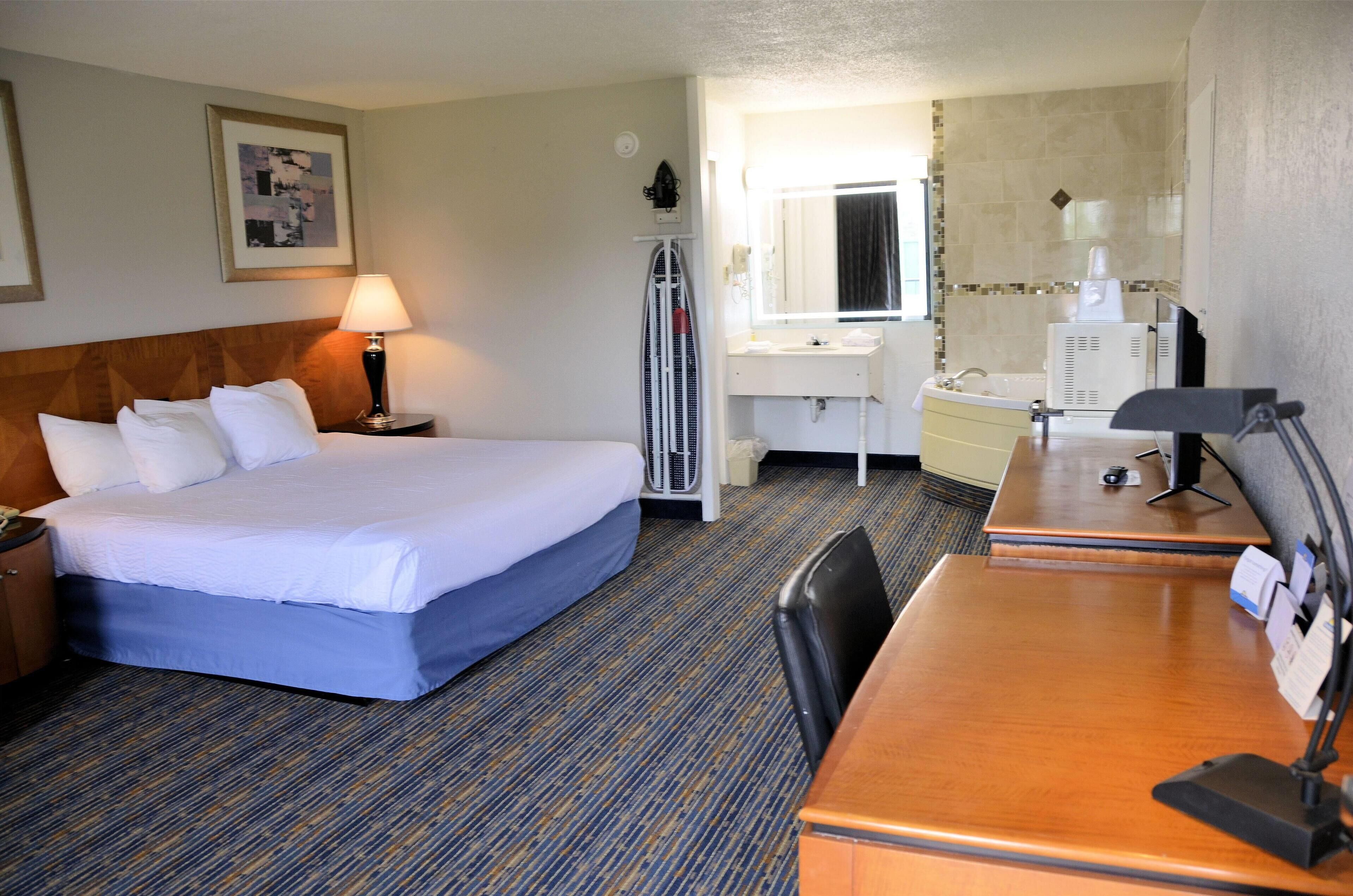 A hotel room with a bed, a desk, a chair, a lamp, a mirror, a window, and a bathroom with a sink and toilet.