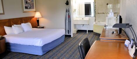 A hotel room with a bed, a desk, a chair, a lamp, a mirror, a window, and a bathroom with a sink and toilet.
