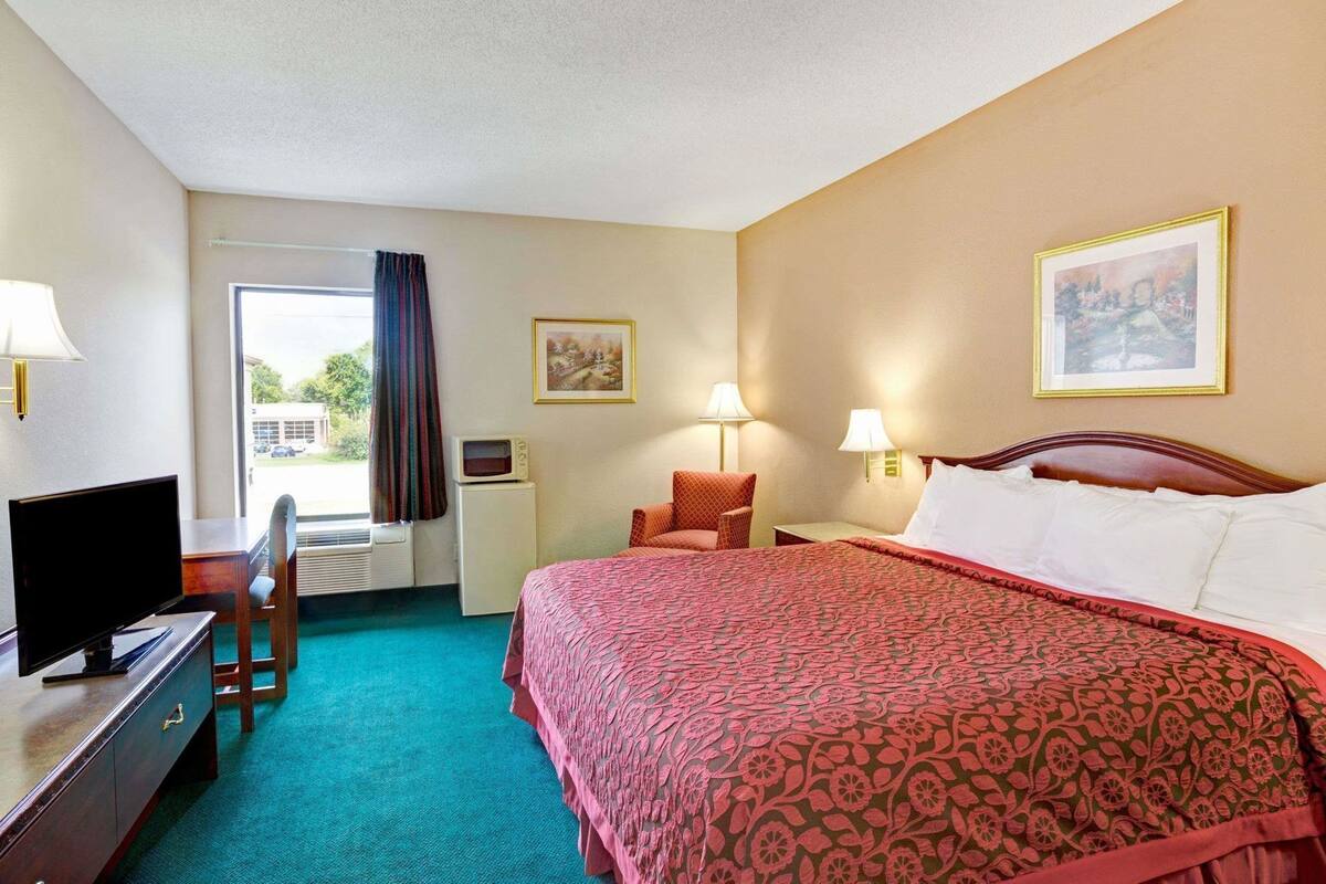 A hotel room with a bed, a desk with a television, a chair, a window with curtains, a wall-mounted lamp, and a framed picture.