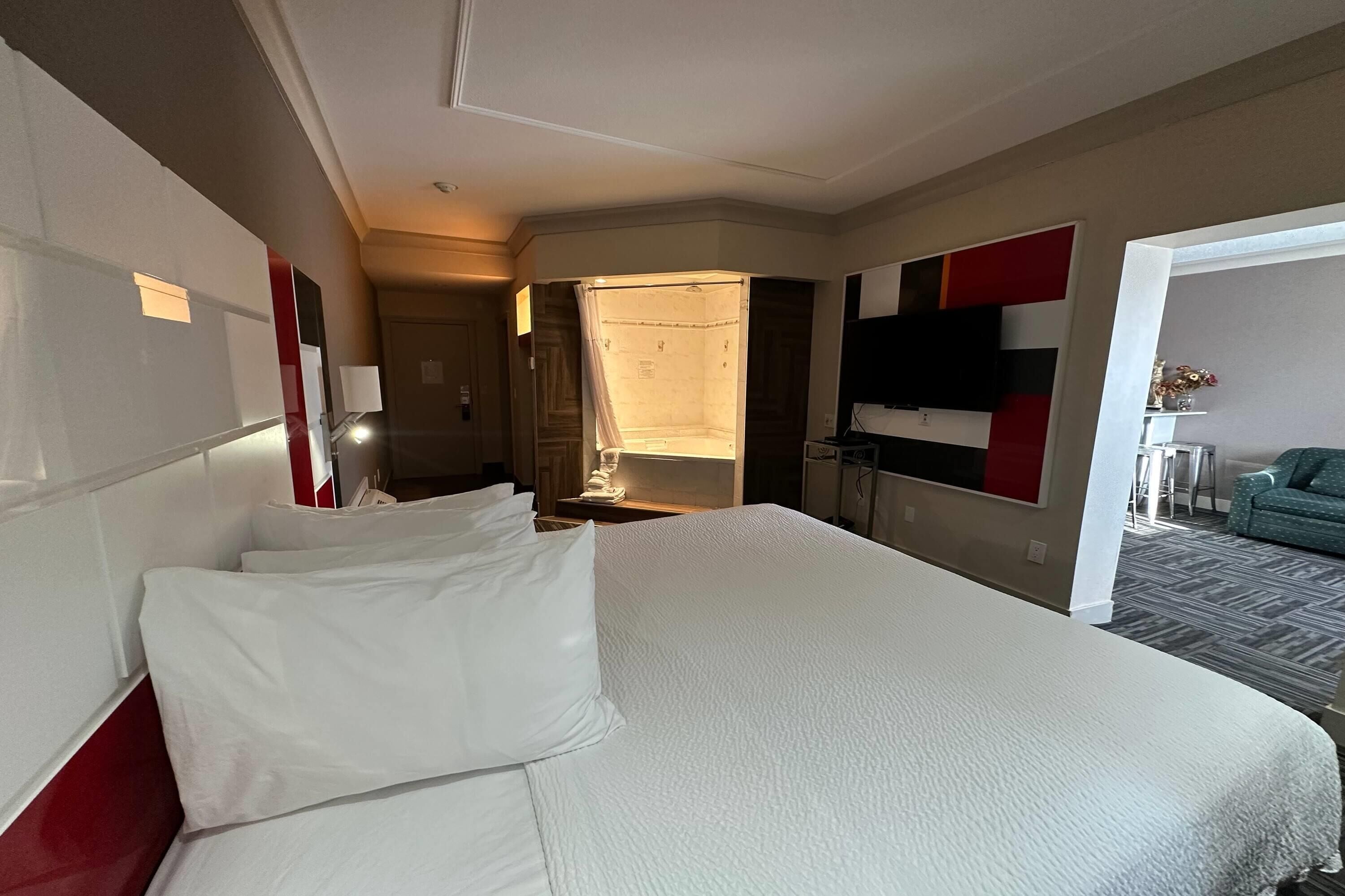A hotel room with a large bed, a desk, a chair, a TV, and a view of the hallway.