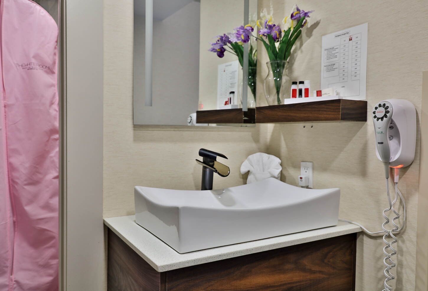A bathroom with a white sink, a towel rack, a mirror, a shelf with toiletries, and a hairdryer.