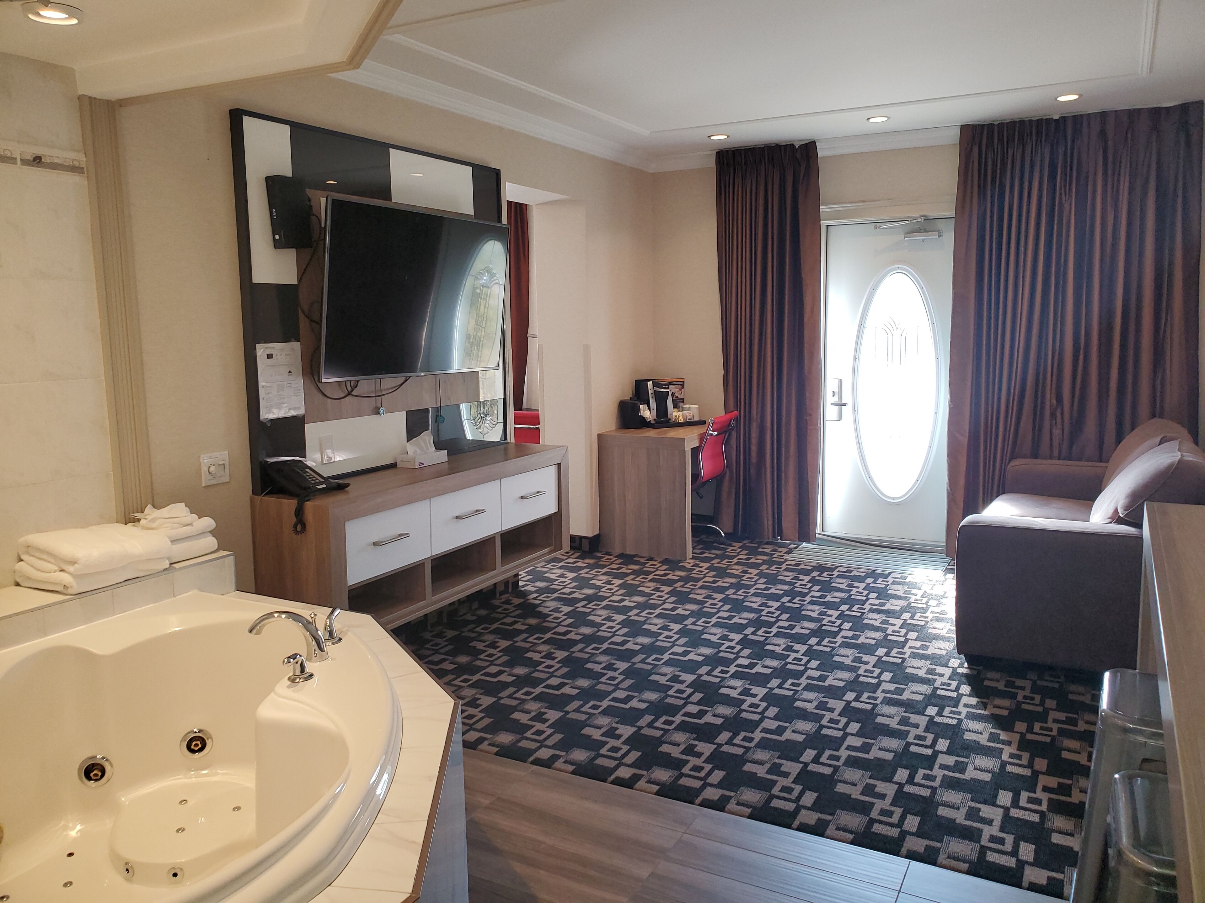 Habitación de hotel con una bañera grande, un escritorio con silla, un televisor y un ventanal con cortinas.