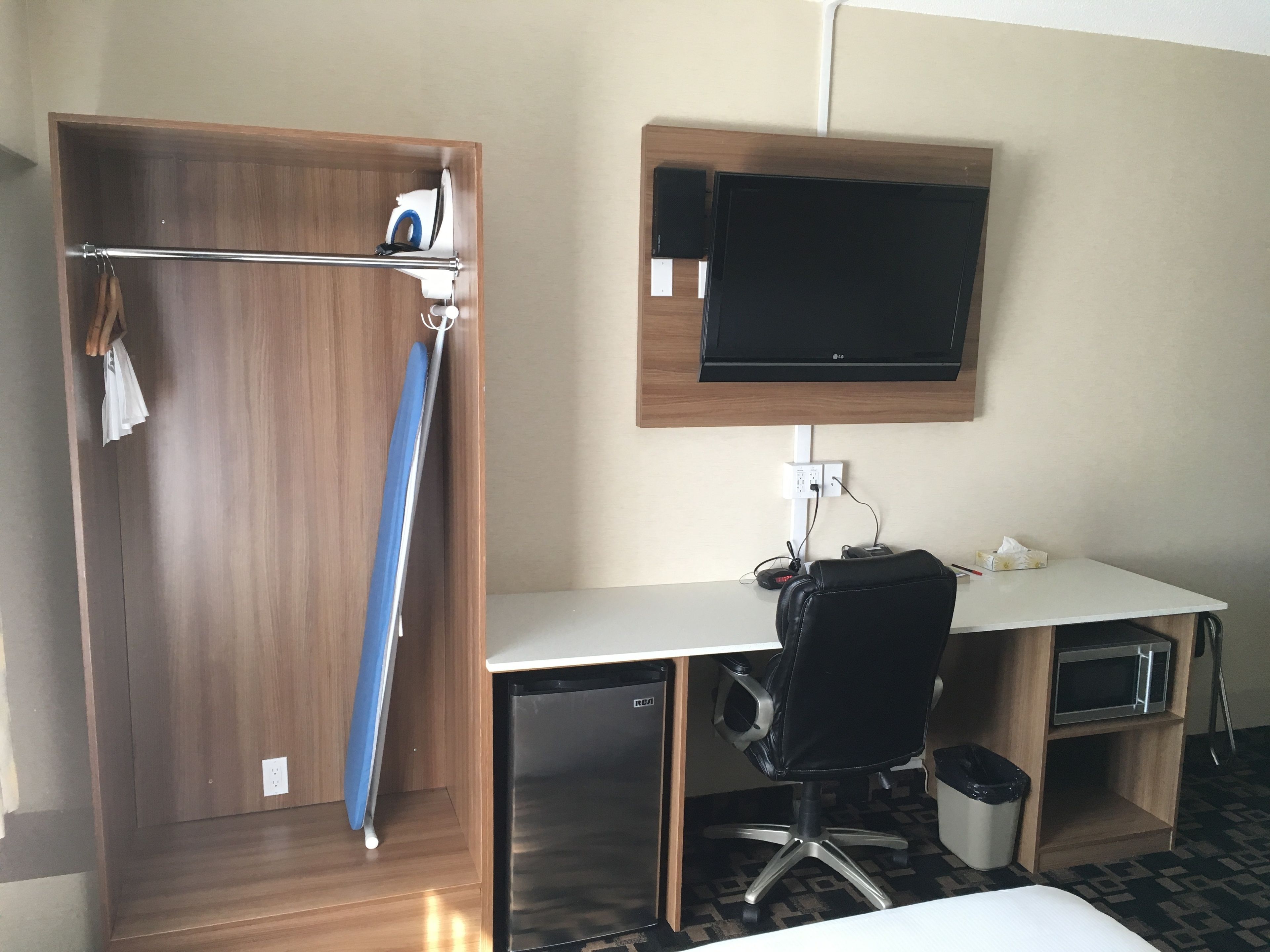 Una habitación de hotel con un armario de madera, un escritorio con una computadora, un televisor montado en la pared y un cesto de basura.