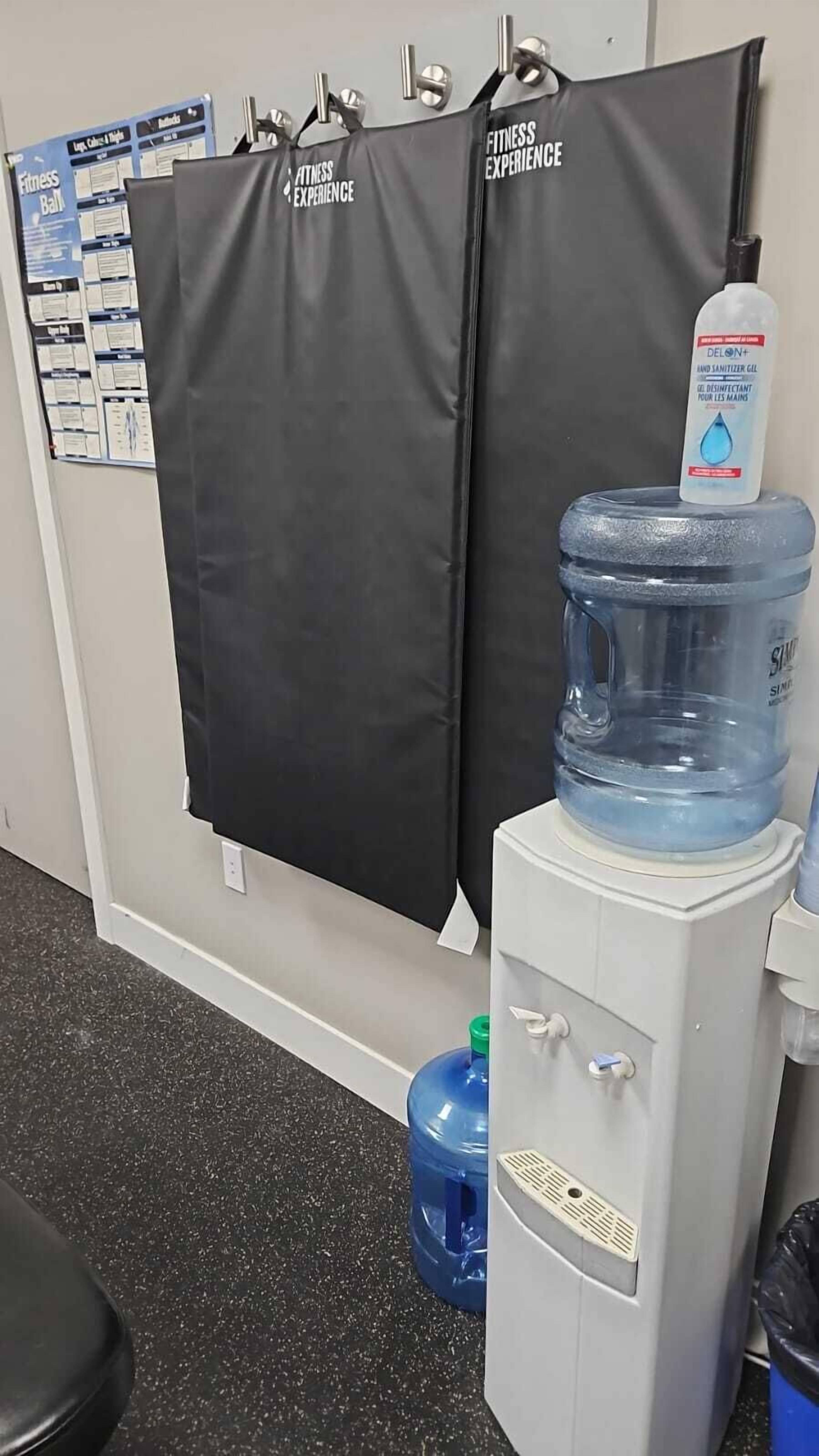 A water dispenser with a bottle of hand sanitizer on top, a large water jug, and two black fitness experience banners on the wall.