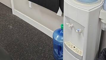 A water dispenser with a bottle of hand sanitizer on top, a large water jug, and two black fitness experience banners on the wall.