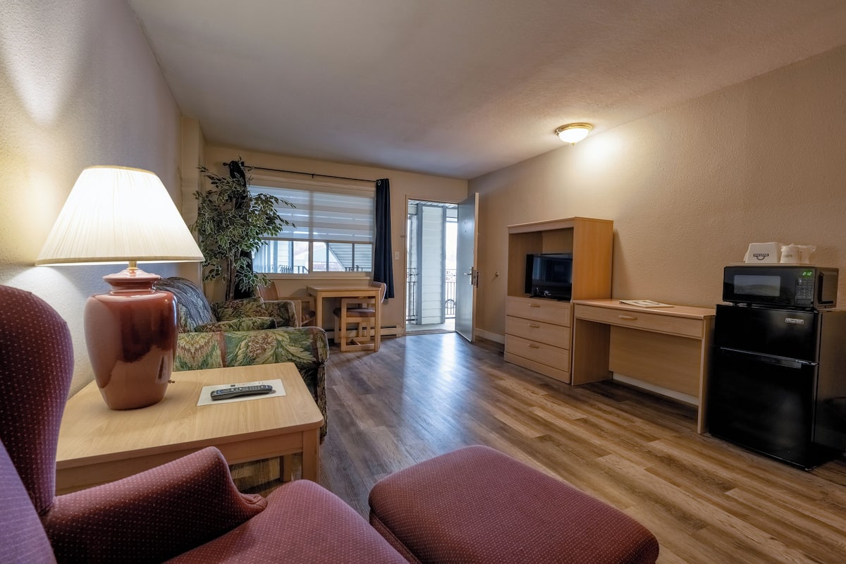 A compact living space with a sofa, a small table with a lamp, a TV, a kitchenette, and a large window.