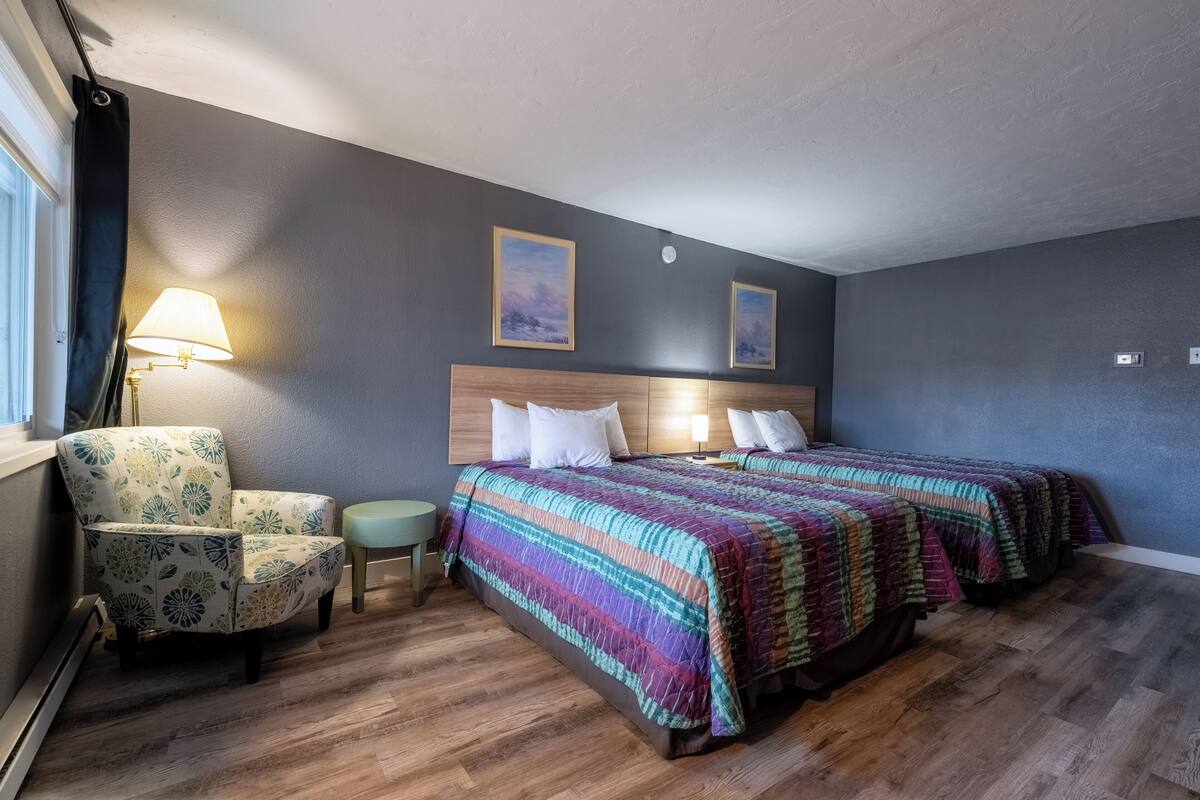 A hotel room with a wooden floor, a bed with a colorful striped bedspread, a chair with a floral pattern, and two wall-mounted lamps.