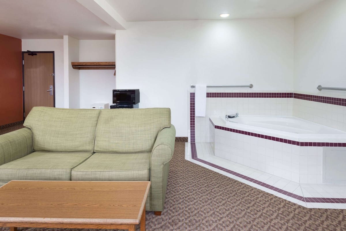 Una habitación con sofá verde, una mesa de madera, una bañera blanca con azulejos rojos y un microondas sobre una mesita.