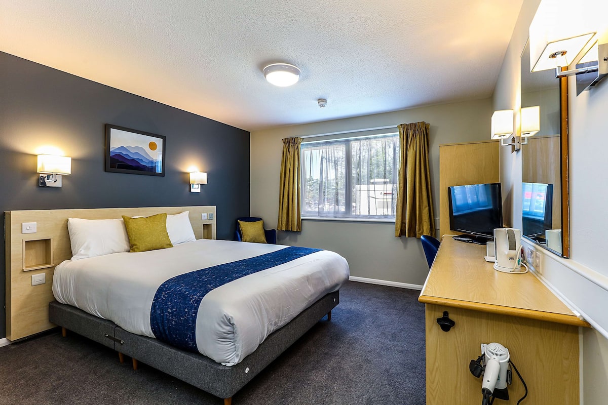 A hotel room with a bed, bedside table, TV, and a window with curtains.