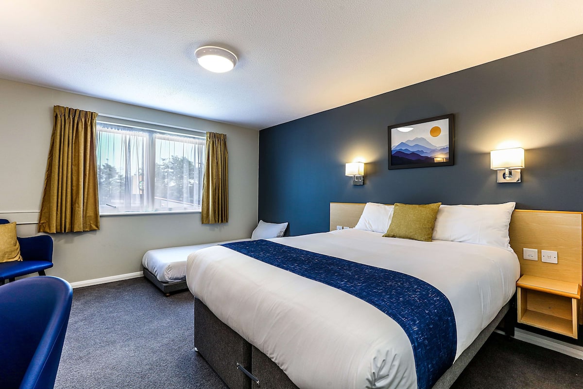 A hotel room with a large bed, a desk, a chair, a window with curtains, and wall-mounted lamps.