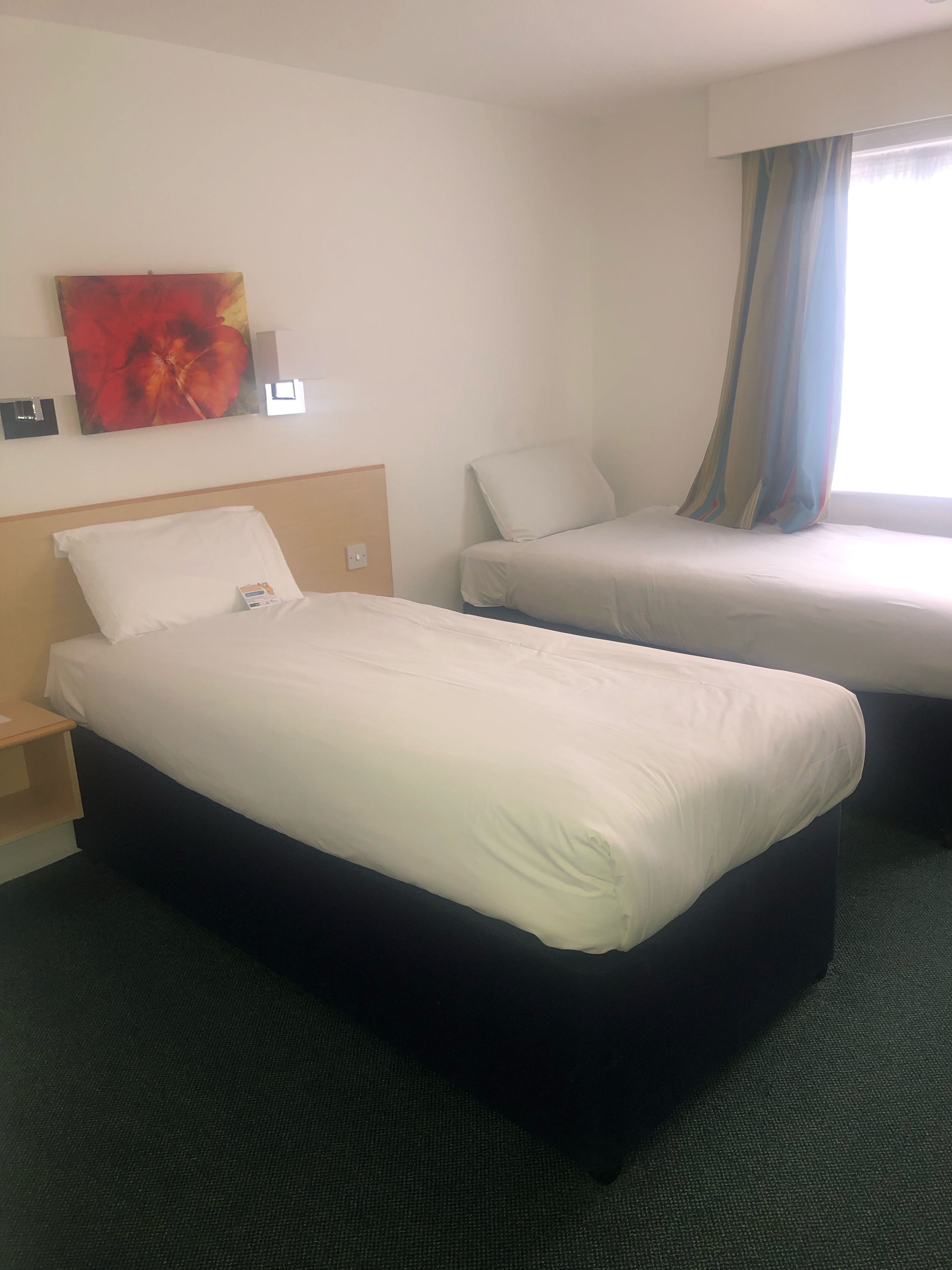 Habitación de hotel con una cama grande, una cama más pequeña, una mesita de noche, una lámpara de pared, un cuadro y una ventana con cortinas.