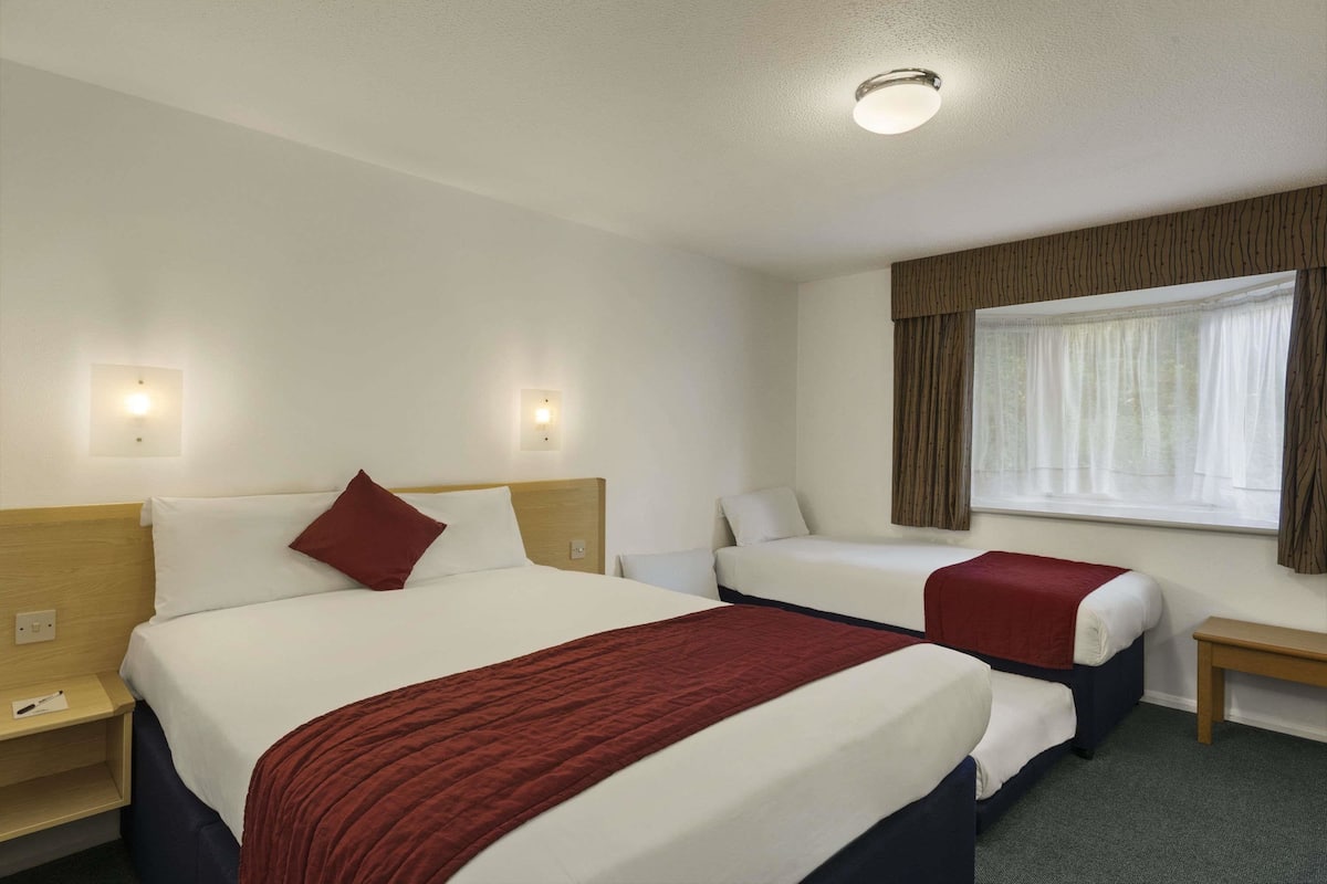 A hotel room with two beds, a wooden headboard, bedside tables, a window with curtains, and wall-mounted lights.