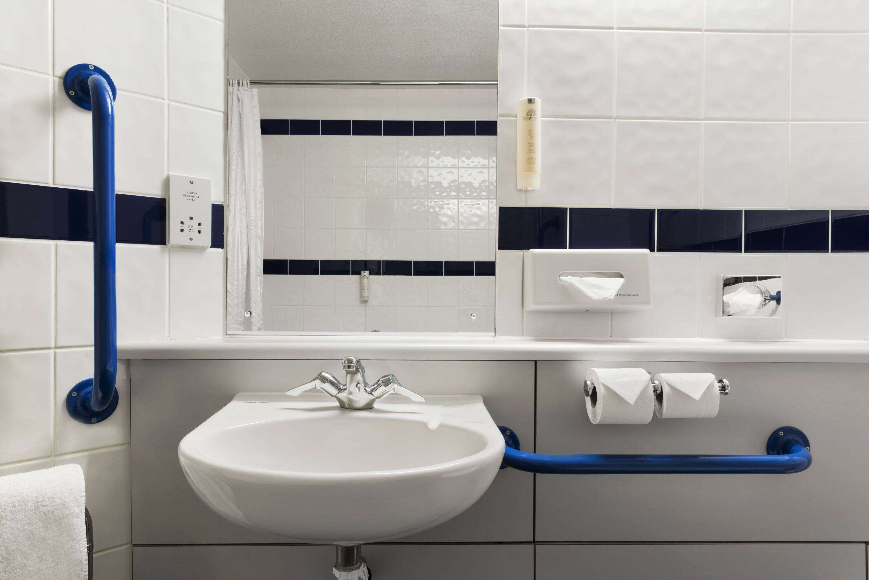 Un baño con un lavamanos blanco, barras de apoyo azules, un espejo y un perchero para toallas.
