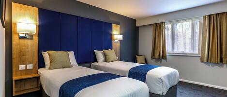 Une chambre d’hôtel avec deux lits, une tête de lit en bois, des lampes fixées au mur, une fenêtre avec des rideaux et un sol recouvert de moquette.
