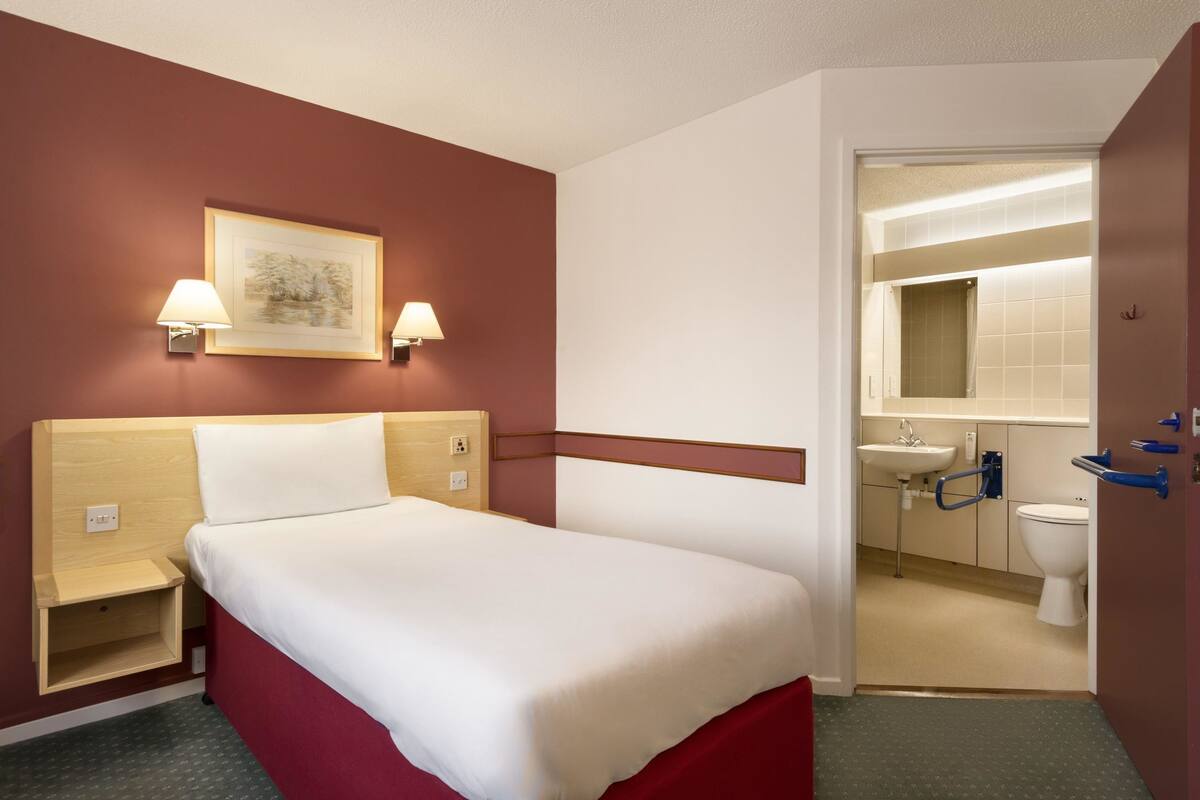 Une chambre d’hôtel comprenant un lit, des tables de chevet, une lampe fixée au mur, un tableau encadré et une salle de bain avec un lavabo et des toilettes.