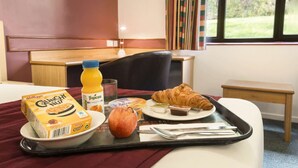 Un plateau de petit-déjeuner avec des céréales, du jus de fruits, une pomme, un croissant et une barre chocolatée, posé sur une table dans une chambre d’hôtel.