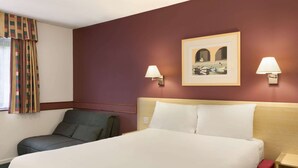 Une chambre d’hôtel avec un lit, deux lampes fixées au mur, un tableau encadré et une chaise.