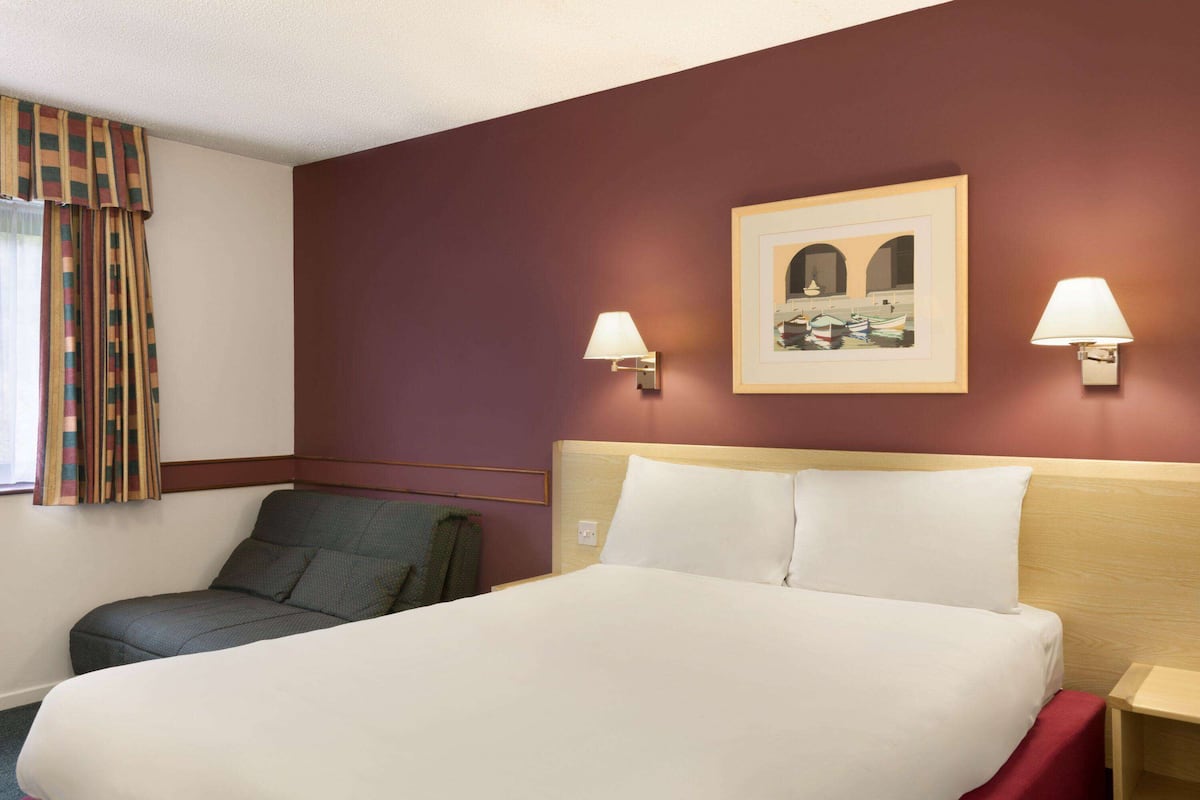 Une chambre d’hôtel avec un lit, deux lampes fixées au mur, un tableau encadré et une chaise.