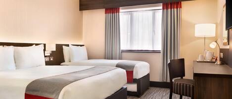A hotel room with two beds, a desk, a chair, a lamp, and a window with curtains.