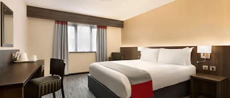 A hotel room with a large bed, a desk, a chair, and a nightstand with a lamp.